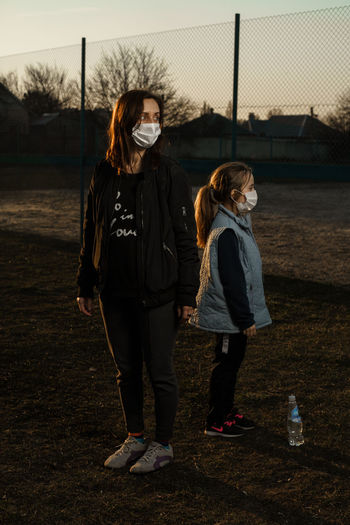 Mother and daughter wearing masks standing on land at sunset