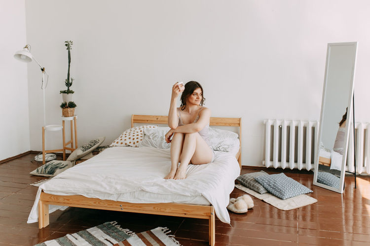 A happy girl in pajamas drinks coffee looks at the phone using technology while lying in bed at home