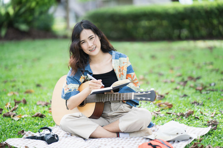 Portrait of young woman writing in diary while holding guitar while sitting at lawn