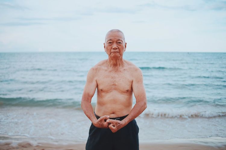 Portrait of shirtless senior man flexing muscles while standing at beach