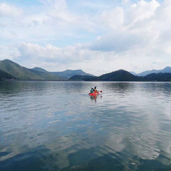 People kayaking in lake against mountains and cloudy sky