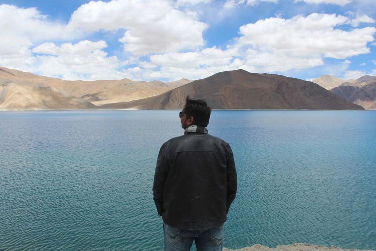 Rear view of man looking at mountains against sky while standing by lake