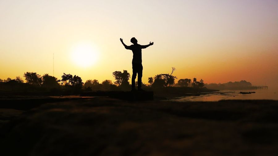 Silhouette man with arms outstretched standing on land against sky during sunset