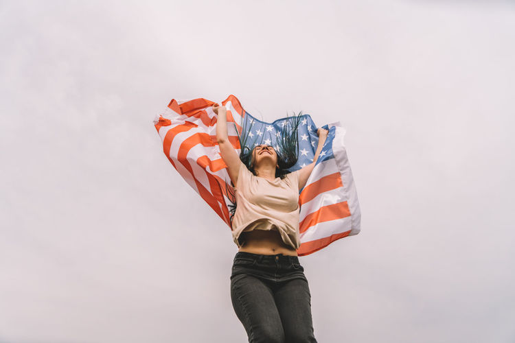Low angel view of young woman holding american flag against sky