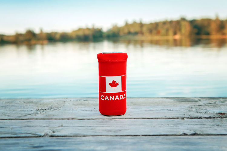 Can of beer in red cozy beer can cooler with canadian flag standing on wooden pier by lake 