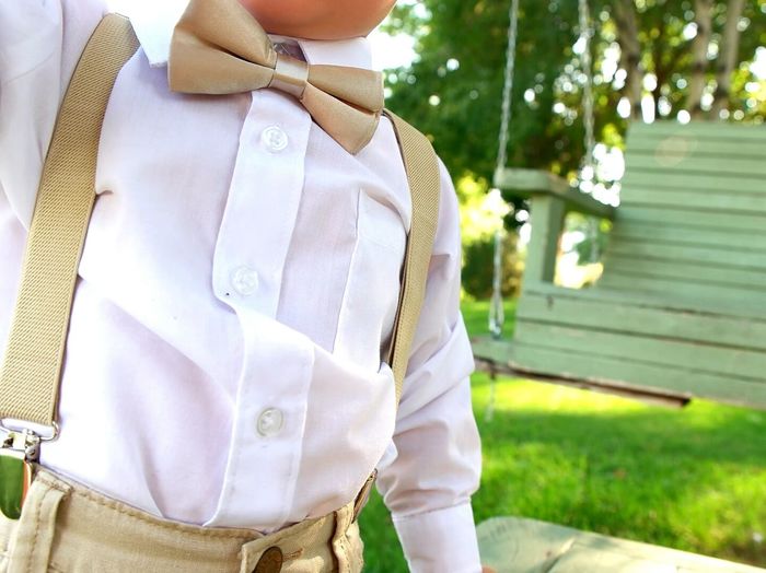 Midsection of boy wearing bow tie and suspenders standing against wooden bench at yard