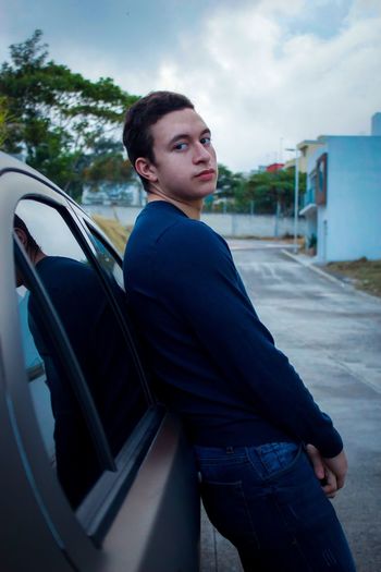 Portrait of teenage boy standing by car against sky