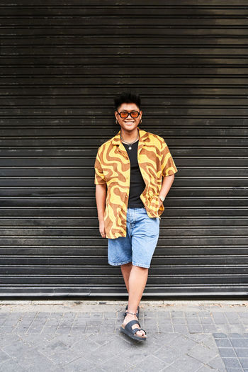 Smiling filipino male in trendy shirt and sunglasses standing at grunge metal wall with hand on pocket and looking at camera with smile
