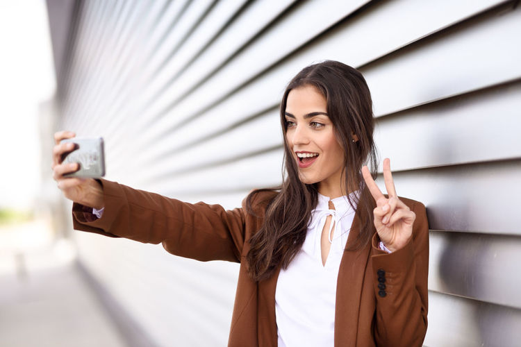 Young businesswoman taking selfie while gesturing peace sign by blinds