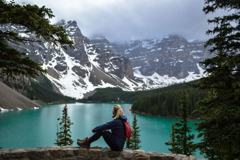 Full length side view of woman sitting by river against mountains during winter