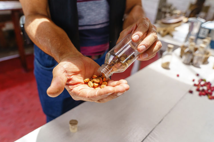 Selective focus of anonymous crop worker pouring raw coffee grains into palm from small plastic bottle in hand above table