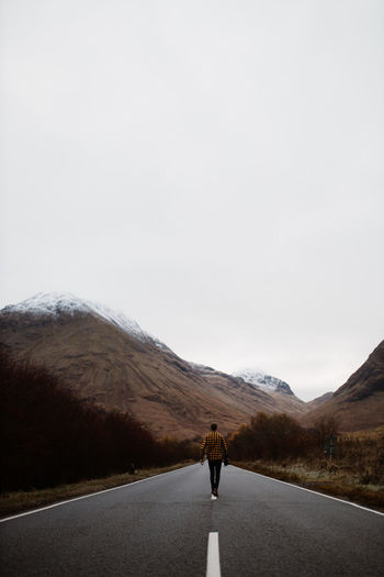 Back view of anonymous traveling male standing on wet empty road and admiring landscape of mountains on cloudy day in scottish highlands