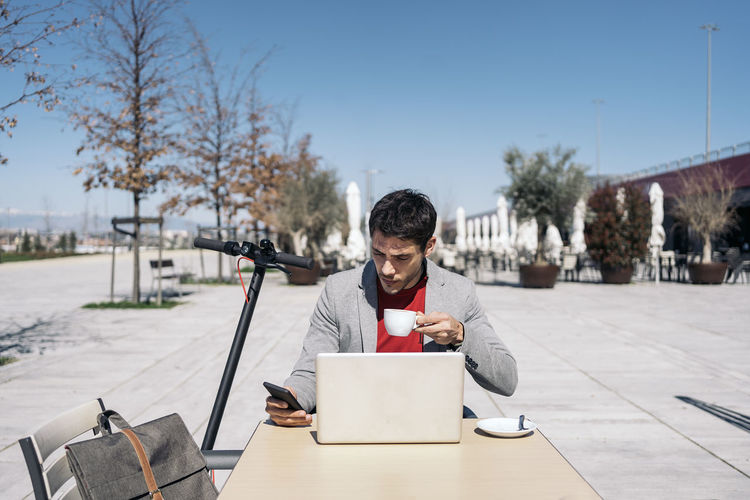 Businessman holding coffee cup while using mobile phone while sitting outdoors