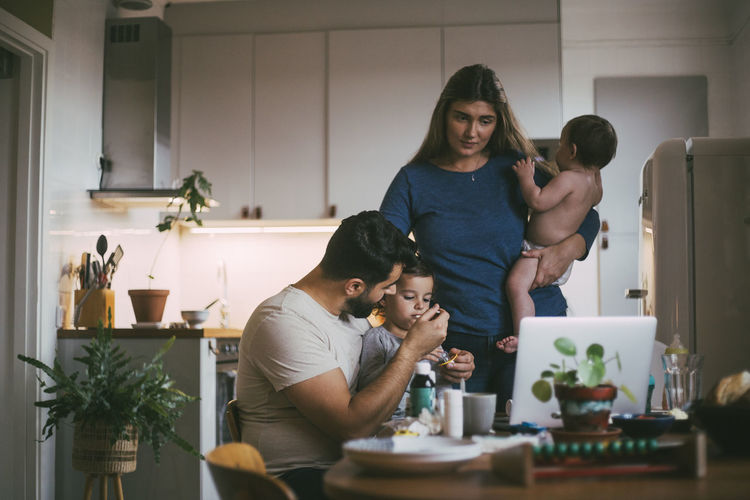 Father feeding son while mother standing with baby girl in kitchen