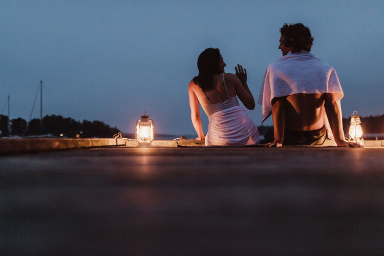 Smiling couple sitting around illuminated lantern on pier against sky