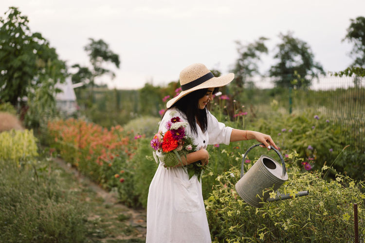 Beautiful brunette girl in a white dress and hat with watering can watering flowers