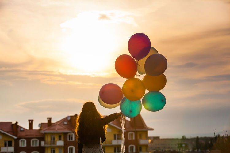Rear view of woman with colorful balloons against sky during sunset