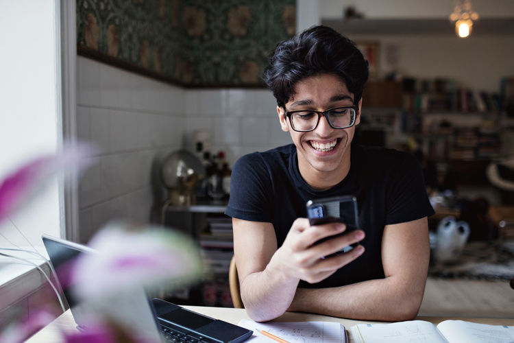 Smiling young man using social media while doing homework at home