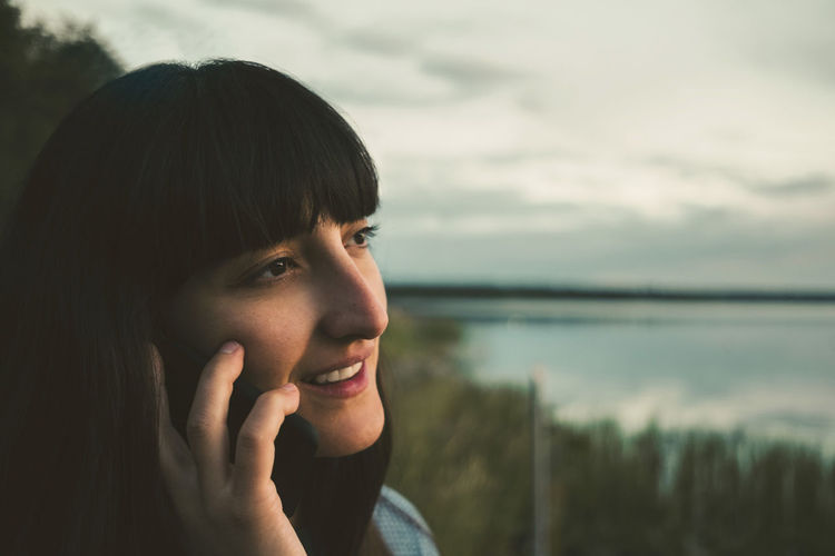 Smiling woman talking on mobile phone by lake against sky