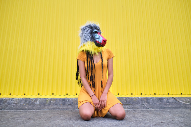 Unrecognizable woman wearing a mandrill mask sitting on her knees on a yellow background.