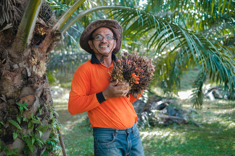Portrait of man wearing hat holding palm fruit standing by tree