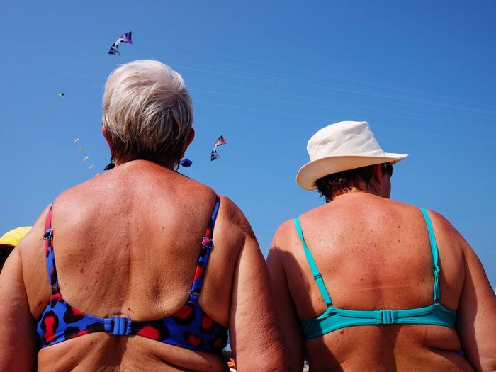 Rear view of female friends wearing bikini tops against kites flying in clear blue sky