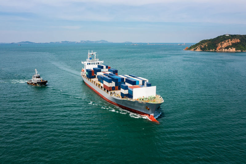 Aerial in front view container ship and tugboat dragging floating in green sea, business 