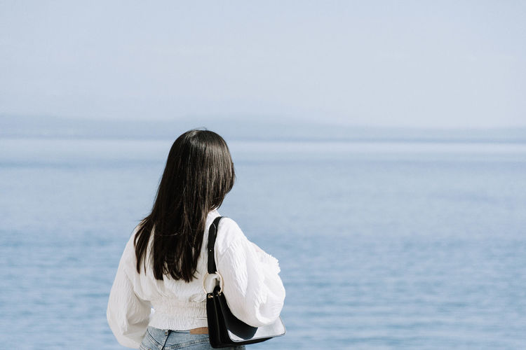 Light and airy rear view of young woman standing on shore of sea.