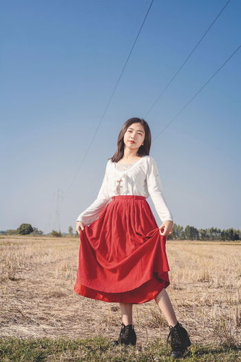 A beautiful woman in a red dress stands among the grasslands and blue sky.