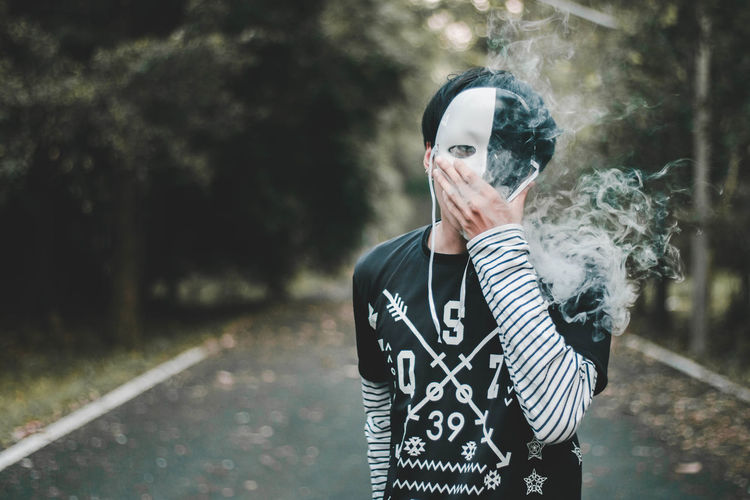 Portrait of man wearing mask while exhaling smoke against trees