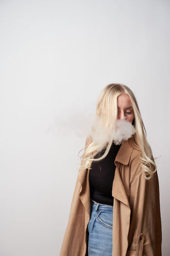 Tranquil female in stylish outfit standing in studio and smoking vape while looking down