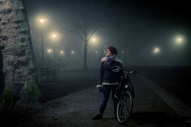 Young woman with bicycle standing on footpath amidst illuminated street lights at park during winter