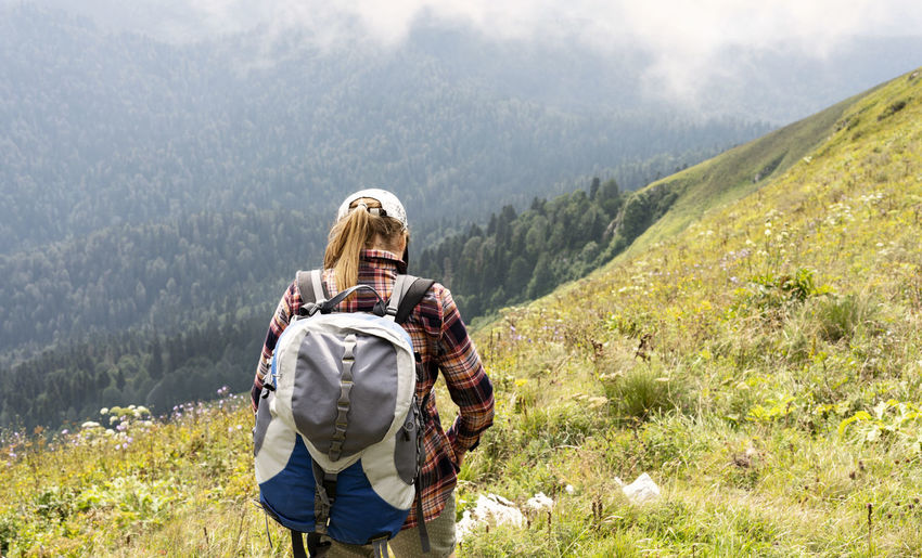  woman in cap and plaid shirt putting on big backpack hiking in green mountains in summer landscape