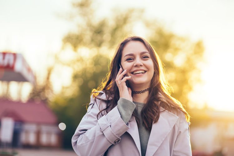 Smiling beautiful girl with long hair in a grey trench coat using smartphone in the spring 