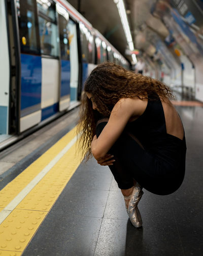 Side view of unrecognizable young ballerina with long curly hair in pointe shoes embracing knees while sitting on haunches in subway station