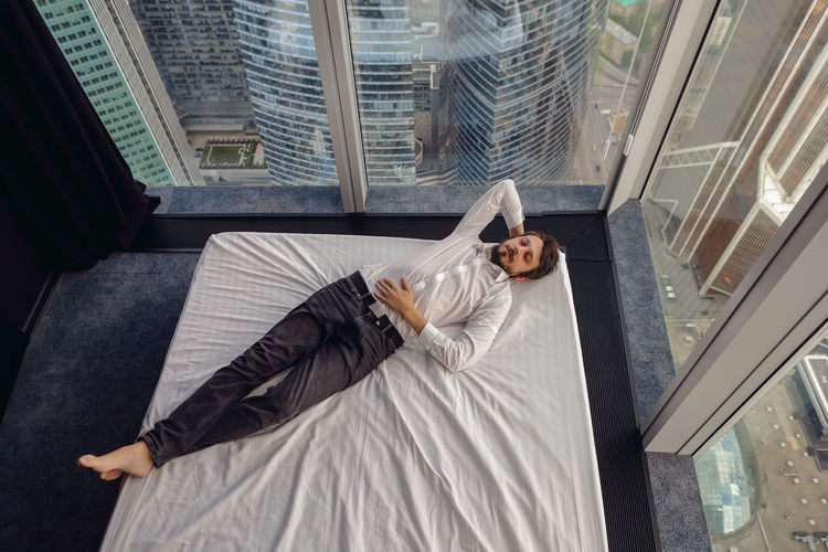 Businessman man in a white shirt is lying on a bed near a large panoramic window resting