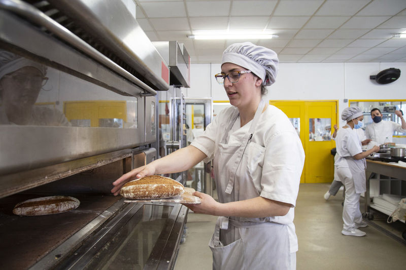 Side view of focused female baker taking out freshly baked bread from hot oven while learning baking in baker school