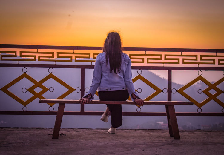 Rear view of woman standing by railing against sky during sunset