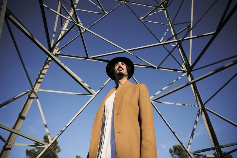 From below of confident young ethnic male in stylish coat and hat looking away while standing against high metal electric tower and blue sky