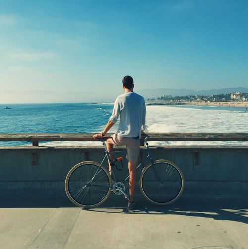 Rear view of man with bicycle by sea against clear sky
