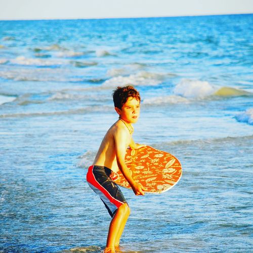 Shirtless boy holding surfboard while standing on shore at beach during sunny day