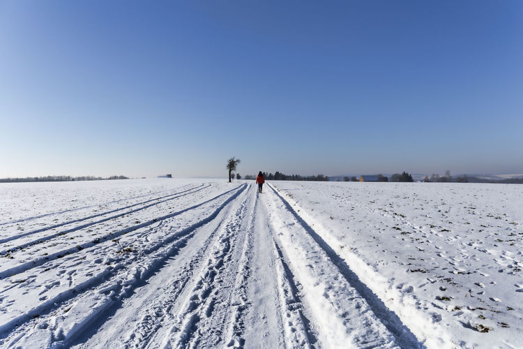 Rear view of person walking on snow covered field against clear blue sky
