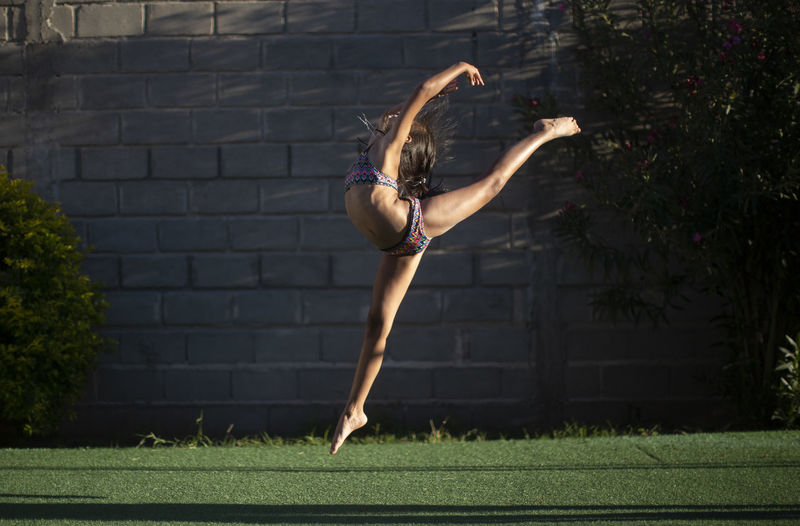 Full length of flexible woman wearing bikini while dancing against wall