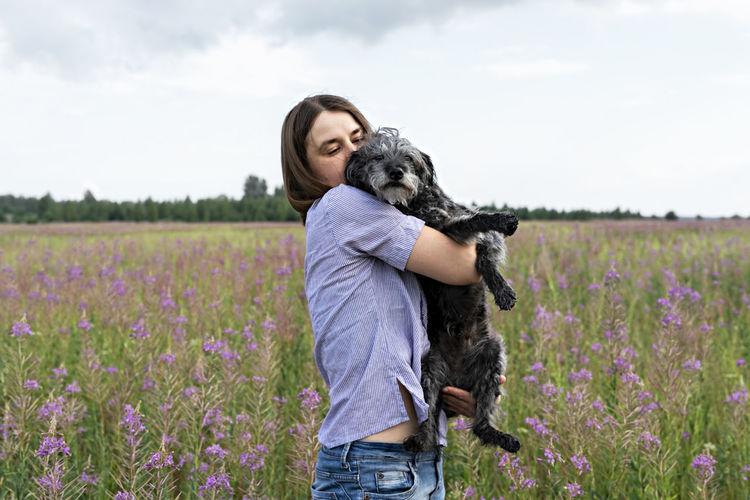 Young woman holding fluffy senior dog in hands on meadow with flowers of fireweed , pet love walking