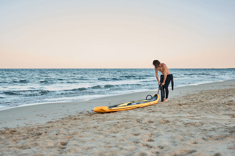 Man in wetsuit inflating paddle board at the seashore on sandy beach