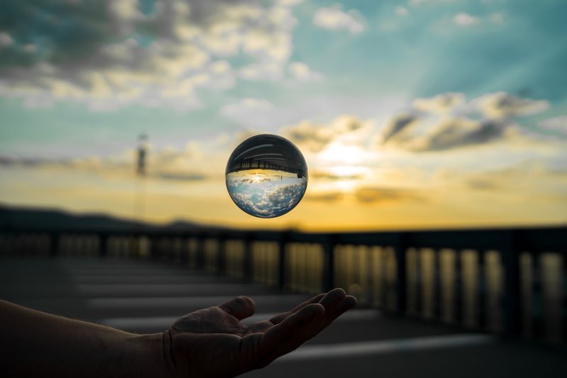 Cropped hand of man catching crystal ball against sky during sunset