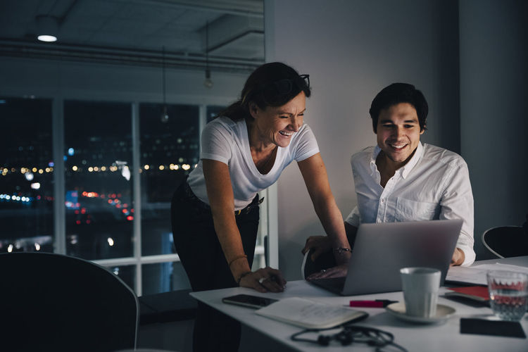 Smiling male and female colleagues discussing over laptop while working late in creative office