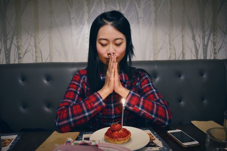 Young birthday woman praying in front of illuminated candle on strawberry tart at restaurant
