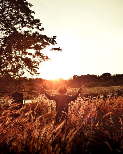 Man with arms raised standing at field against sky during sunset