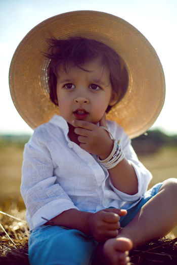 Baby boy in straw hat and blue pants sitting on a haystack in a field in autumn